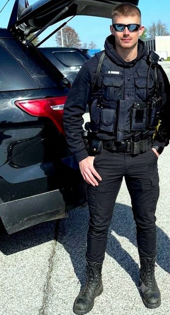 A young man with a buzzcut and sunglasses poses in a full black tactical uniform with a load-bearing vest and combat boots next to a vehicle.