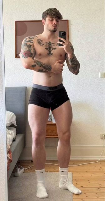 A tattooed, muscular young man poses for a mirror selfie wearing black boxer briefs and white crew socks in a bedroom.