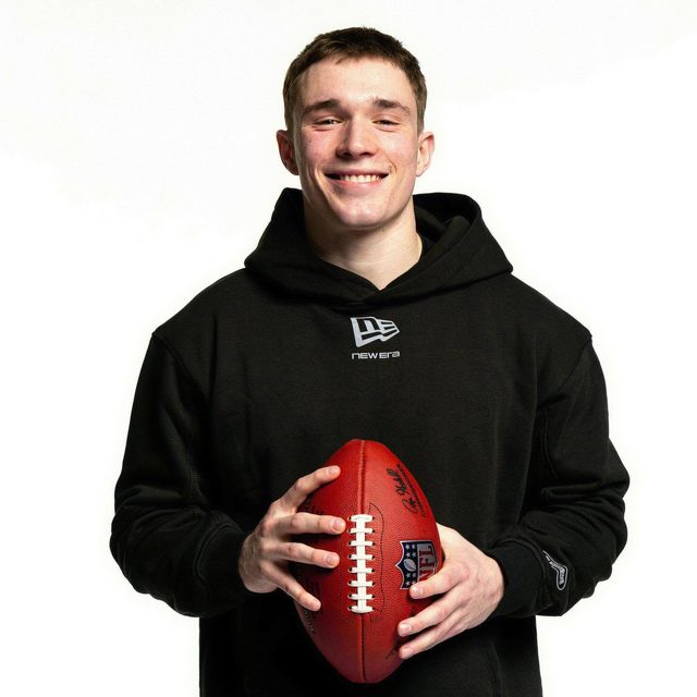 A smiling young man with a short haircut wears a black New Era hoodie while holding an NFL football against a white studio background.