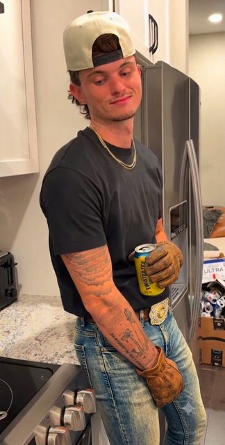 A tattooed young man wearing a backwards baseball cap, denim jeans, and brown leather work gloves stands in a kitchen.