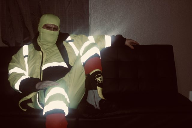 A man sits on a dark couch wearing a full high-visibility construction uniform and a matching balaclava that hides his face.