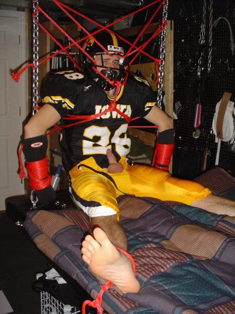 A man in a football uniform is suspended and restrained with red ropes on a padded surface.