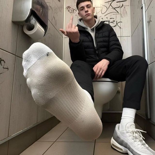 A young man sits on a toilet in a public restroom with one socked foot extended.