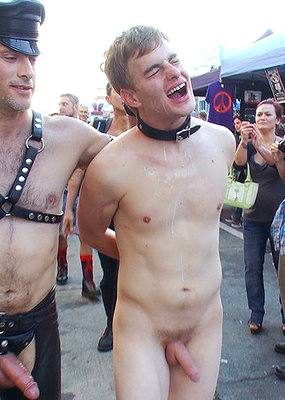 A naked young man wearing a black leather collar stands outdoors at a leather street festival beside a man wearing a muir cap and chest harness.