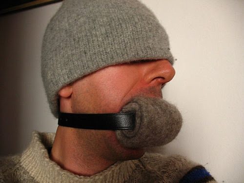 A man wears a fabric gag secured with a leather strap and a grey beanie indoors.