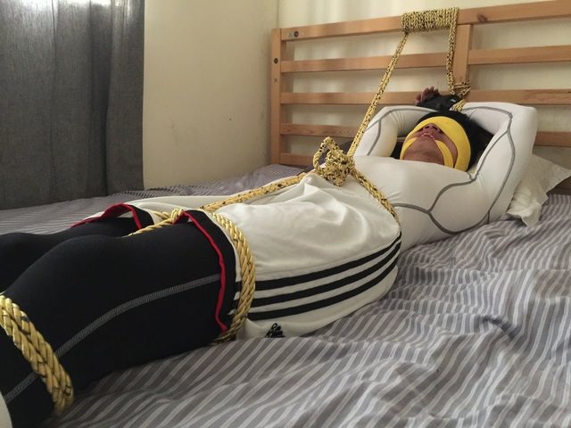 A man wearing sportswear is tied with gold rope and blindfolded on a bed in a bedroom.