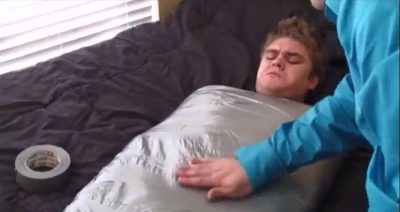 A man is wrapped in plastic film and tape on a bed while another person touches him.