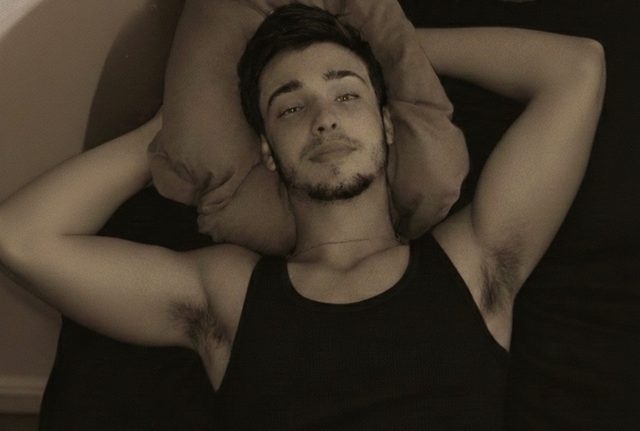 A young man with a light beard and hairy armpits lies on a pillow in a black tank top, looking up at the camera.