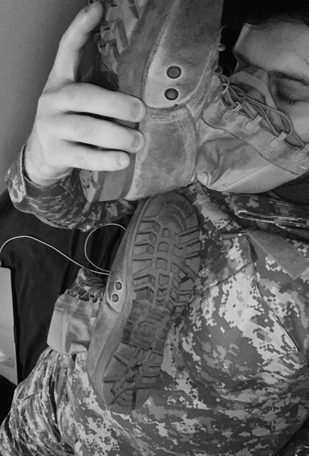 A man in a military camouflage uniform presses the sole of a combat boot against his face.