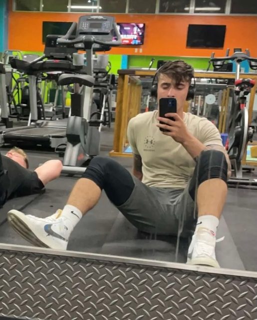 A man in sportswear sits on the gym floor taking a selfie, with another person visible nearby.