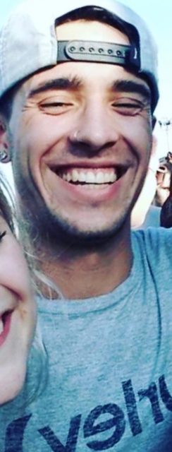 A young man in a cap with a nose piercing smiles broadly outdoors.