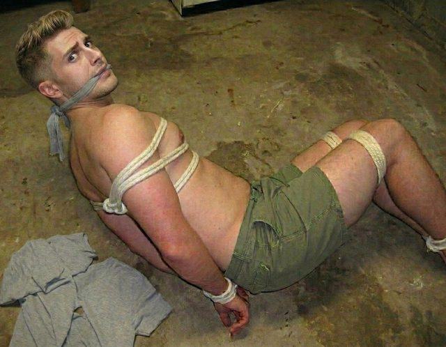 A muscular man is restrained with rope and a cloth gag on a concrete floor.