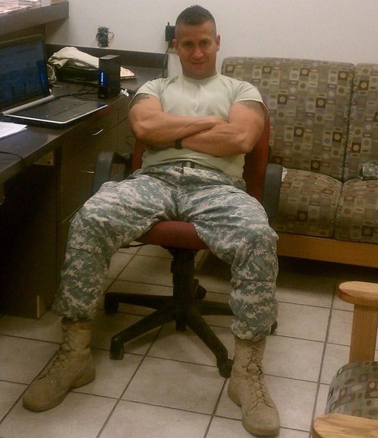A muscular man in a military uniform and boots sits in an office chair.