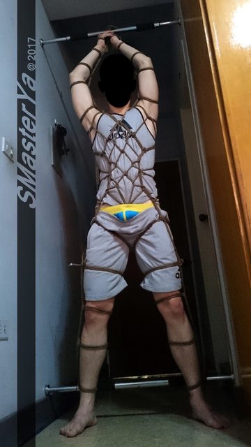 A man is suspended by his bound hands to a pull-up bar while secured in full body rope bondage.