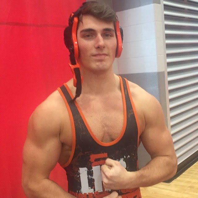 A muscular man in a wrestling singlet and headgear poses in a gym.