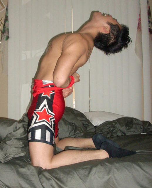 A man in wrestling shorts and socks kneels on a bed with his wrists restrained.