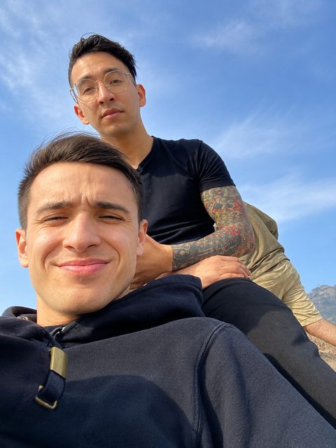Two clean-shaven men, one with a full sleeve tattoo, pose for a selfie while outdoors in a mountainous area.