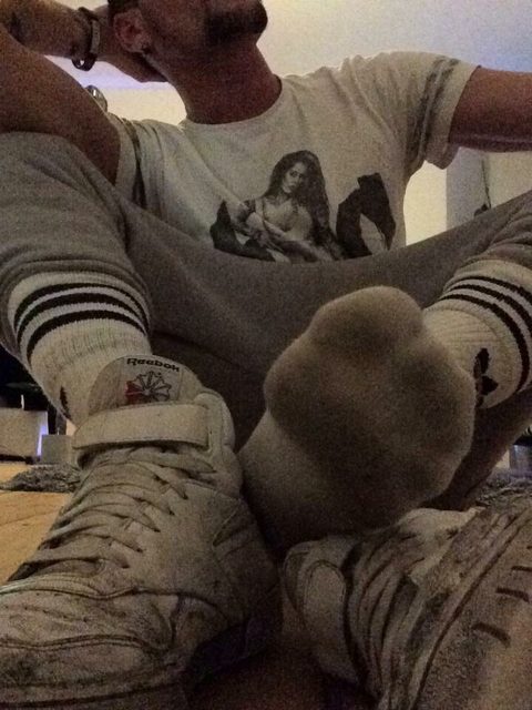 A man in a t-shirt, sweatpants, socks, and sneakers is lounging indoors.