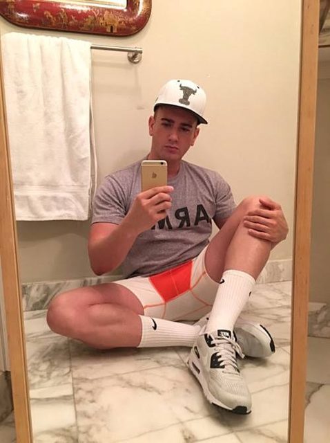 A muscular man in sportswear sits on a marble bathroom floor taking a mirror selfie.