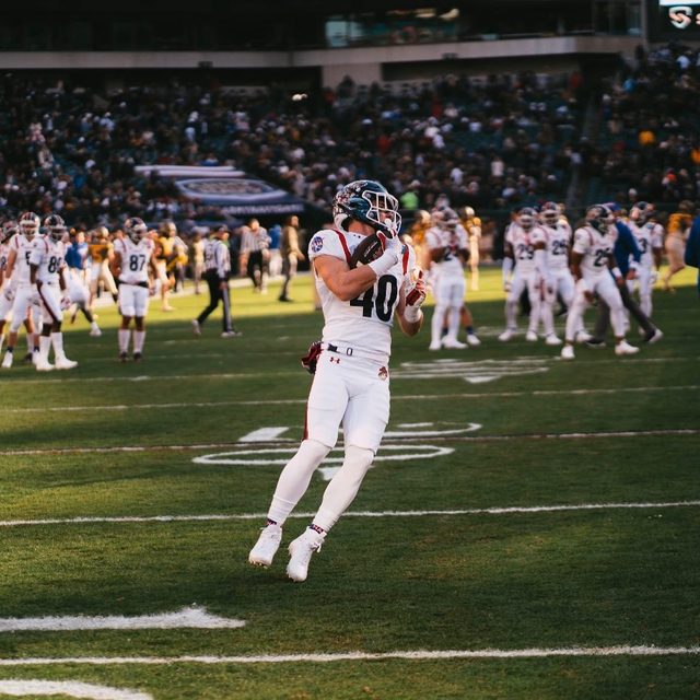 A muscular football player in a full white uniform catches a ball on a stadium field.