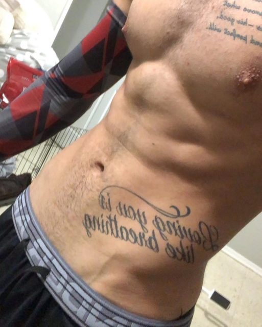 A muscular man with tattoos on his chest and torso wears sportswear arm sleeves and athletic shorts with underwear peeking out.