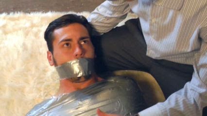 A man is gagged and restrained with duct tape by another person indoors.