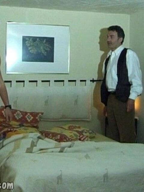 A mature man dressed in a white shirt and black vest stands in a bedroom beside a bed.
