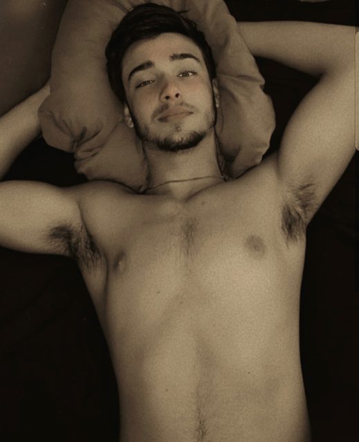 A shirtless man with a beard and chest hair lies on a bed.