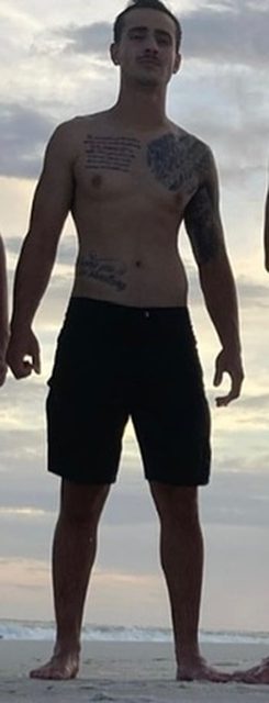 A man with tattoos stands on a beach in black swim shorts with the ocean in the background.