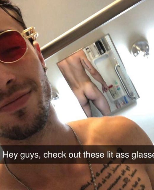 A man with sunglasses and tattoos takes a selfie, reflecting his nude body in a bathroom mirror.