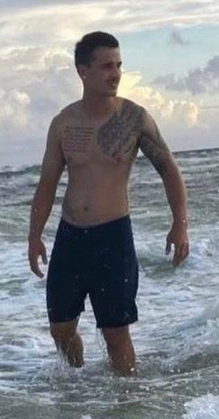 A man with tattoos stands in the ocean waves on a cloudy day.