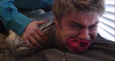 A man with a red gag is restrained with plastic wrap while another person presses on his back.