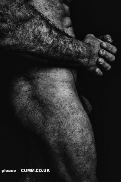 A close-up black and white photograph depicting the hairy torso and thigh of a mature man standing with clasped hands.