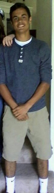 A young, lean man stands indoors wearing a blue henley, khaki shorts, and white Nike tube socks.