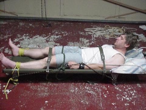 A man is lying on a stretcher, restrained with ropes and straps.