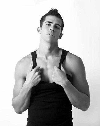 A black and white portrait of a smooth, athletic young man pulling on the straps of his black tank top against a plain studio backdrop.
