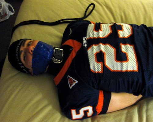 A man wearing a jersey, collar, and a blue tape gag is restrained on a bed.
