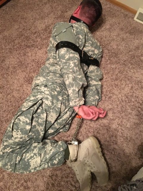 A person in military uniform is restrained on a carpeted floor, wearing a collar and mask.