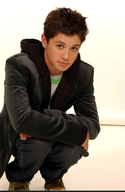 A young man crouches in a studio wearing a jacket, hoodie, and jeans.