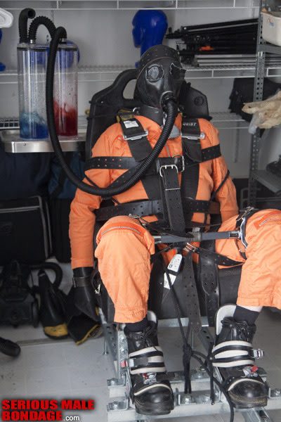 A person in an orange jumpsuit, mask, and harness is restrained in a specialized chair with medical equipment nearby.