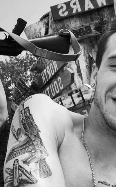 A smiling man with a weapon tattoo poses outdoors near military-themed signs.