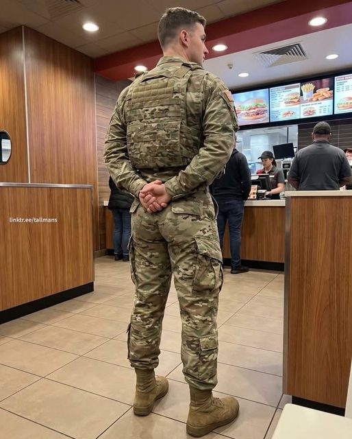 A man with an athletic build stands in a public fast-food restaurant wearing a full military camouflage uniform and tactical vest.