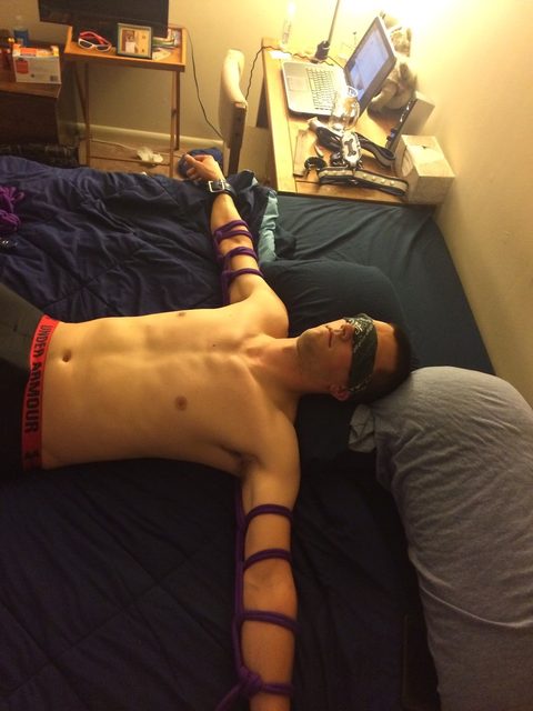 A man is tied with rope and blindfolded on a bed in a bedroom.