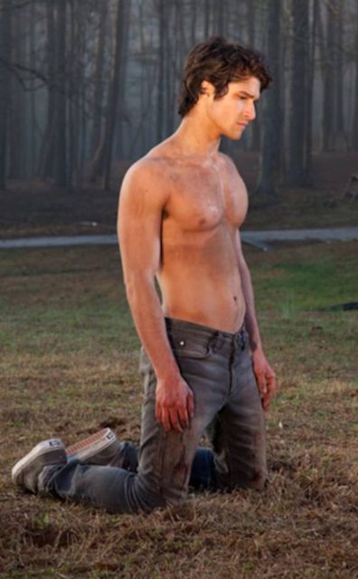 A shirtless young man kneels in dirty jeans and sneakers on grassy ground in an outdoor setting.