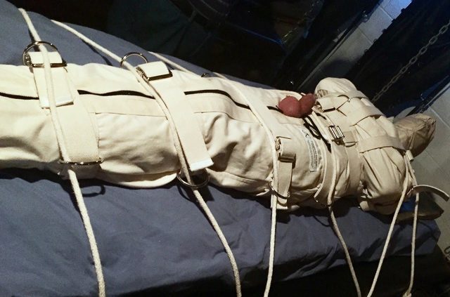 A person is tightly restrained in a straitjacket and ropes on a bed.