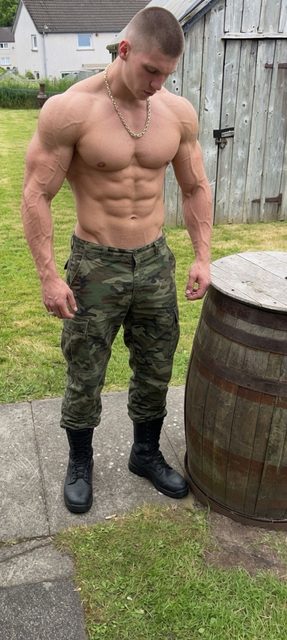 A shirtless muscular man with a buzzcut stands in a backyard wearing camouflage pants and combat boots.