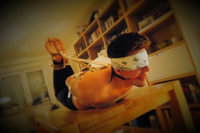 A man is tied with rope, blindfolded, and has a mouth spreader while lying on a table.