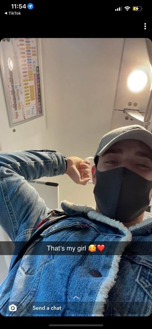 A person in a denim jacket, cap, and face mask takes a selfie while flexing their arm.