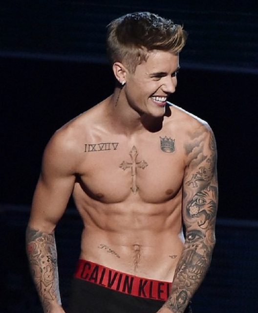 A shirtless, tattooed young man with defined abs and chest smiles while wearing low-slung Calvin Klein underwear.