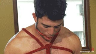 A muscular man is tied with red rope bondage and has a gag in his mouth.